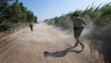 U.S. Border Patrol agents run down a dirt road amid swirling dust stirred up by a helicopter during a search for people who had crossed into the United States illegally and were hiding in heavily wooded areas and sugar cane fields near McAllen, Texas. Photo by Mike DuBose, UMNS.