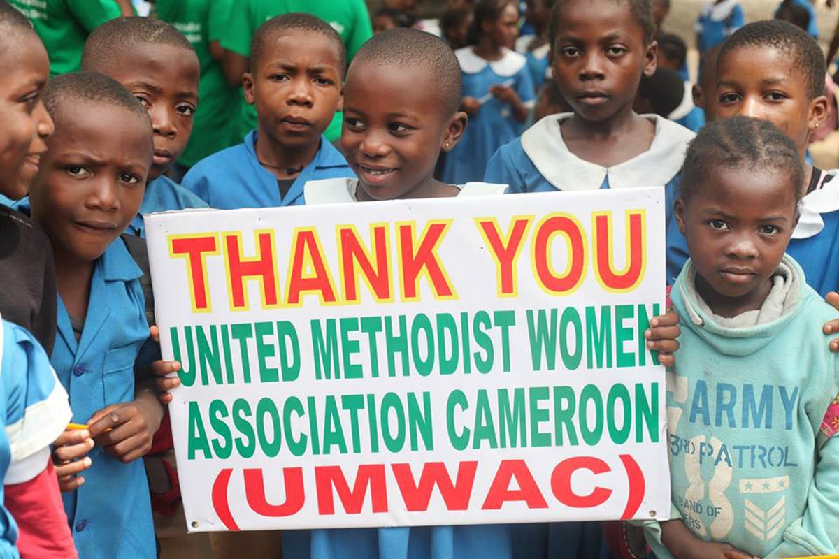 Children gather behind a poster featuring the motto of the United Methodist Women Association Cameroon: “Faith-Love-Action.” Photo by Marius Bonfeu.