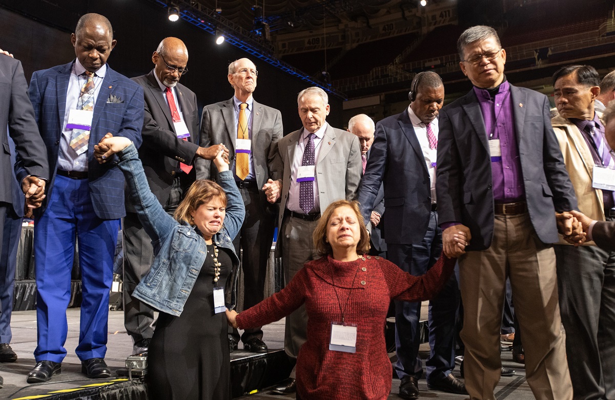 Os delegados da Flórida, Rachael Sumner (à esquerda) e a Revda. Jacqueline Leveron (frente à direita) da Conferência da Flórida, juntam-se em oração com bispos e outros delegados na frente do palco antes de uma votação chave sobre as políticas da igreja a. Foto de Mike DuBose, UMNS.