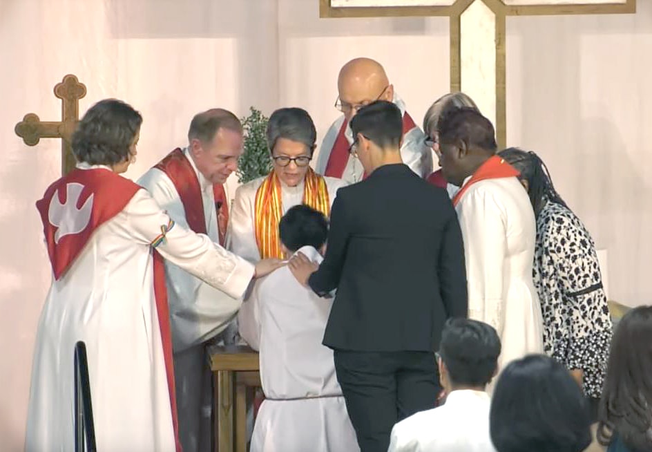 Northern Illinois Bishop Sally Dyck lays hands on the Rev. M Barclay, ordaining Barclay as a deacon in full connection. The cabinet and Barclay’s partner and sponsor, the Rev. Anna Blaedel, also surround the new deacon to pray. Video image courtesy of Northern Illinois Conference livestream.