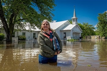 Shelli Pleasant, pastora da Igreja Metodista Unida Avant, fica em frente à igreja com água até as coxas, um dia depois que uma série de tempestades em 20 de maio resultou em enchentes quase históricas da cidade. Foto de John Pleasant, Conferência Anual de Oklahoma.