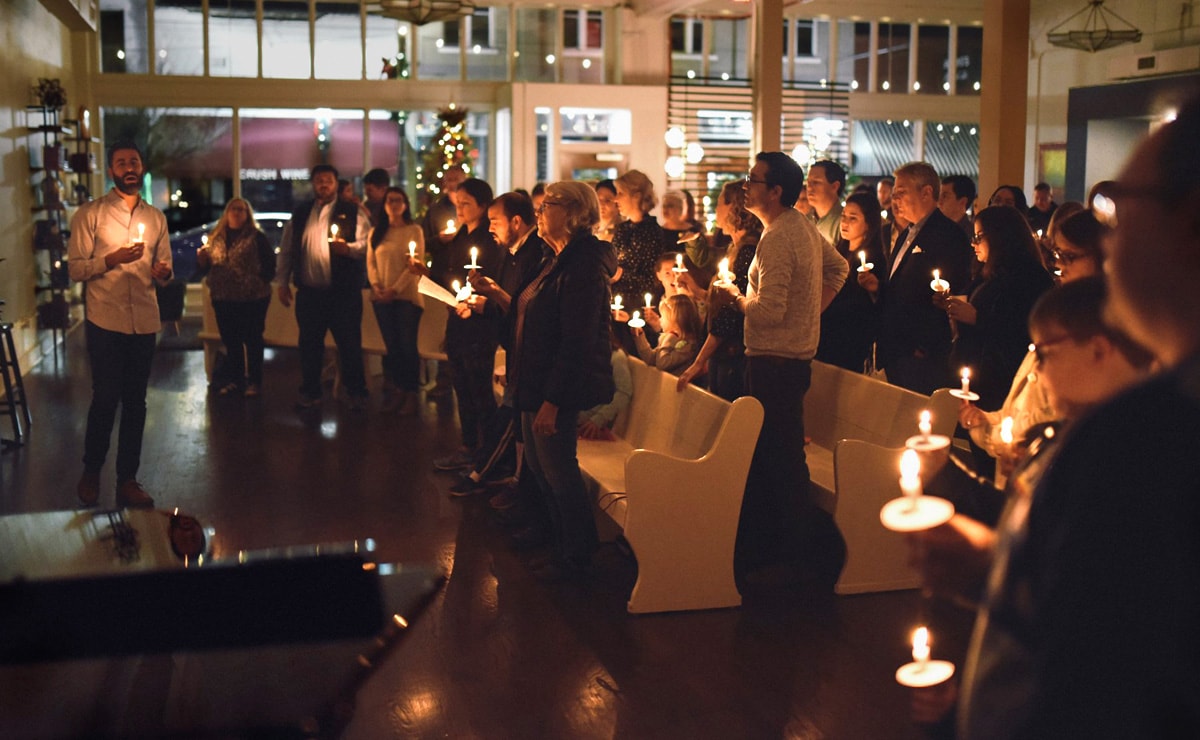 El Rev. Will Choate, a la izquierda, ayuda a los fieles a cantar "Noche de Paz" a la luz de las velas en la víspera de la Navidad de 2018 en la Iglesia Metodista Unida (IMU) Argenta en North Little Rock, Arkansas. La creciente congregación y otros/as en toda la conexión metodista unida se están preparando para hacer que los/as visitantes se sientan bienvenidos/as. Foto cortesía de la IMU Argenta.