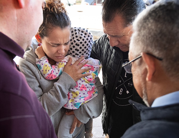 The Rev. Arturo Gonzélez Sandouzl (second from right) and other members of the United Methodist Immigration Task Force pray with Isabél and her 16-month-old daughter Kassandra at a makeshift camp near the bridge leading to the U.S. in Matamoros, Mexico. The mother and daughter traveled from Nicaragua in hopes of seeking asylum in the U.S. Photo by Mike DuBose, UM News.