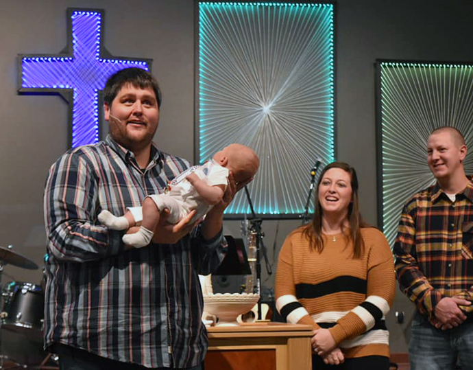 El Rev. Jason Martens (izquierda) sostiene a un bebé durante un bautismo en la IMU Celebration (La Celebración) en Brandon, Dakota del Sur. Este es el segundo proceso de renacimiento de iglesias que lidera Martens. Durante su permanencia en la IMU en Salem, Dakota del Sur, la asistencia aumentó de 25 a 115. Foto cortesía de la página de Facebook de la IMU Celebration.