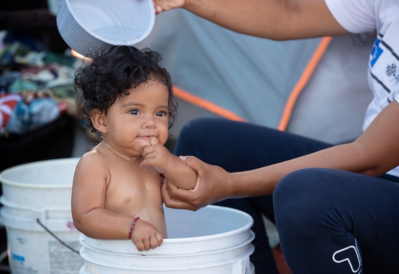 Kelani, de ocho meses, cuya madre huyó de la violencia de las pandillas en Honduras, se baña en un balde de plástico en un campamento improvisado para migrantes en el cruce fronterizo de El Chaparral en Tijuana, México. Foto de Mike DuBose, Noticias MU.