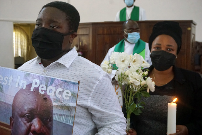 Nyasha Buta holds a photo of his father who died from COVID-19 during a service at Chisipiti United Methodist Church in Zimbabwe honoring those who passed from the disease but were not given a normal funeral due to restrictions. His mother, Anne Buta, stands behind him holding flowers and a candle. The Rev. Daniel Mutidzawanda stands in the background between them. Photo by Kudzai Chingwe, UM News.