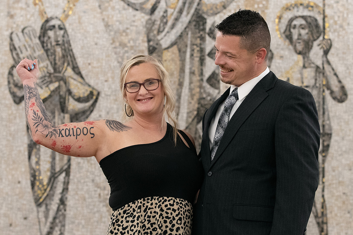 Amber Blankenship shows off a tattoo honoring Kairos Prison Ministry, a program which she credits with changing her life while she was incarcerated at the Lakin Correctional Center in West Columbia, W.Va. “I told God that if he would heal my broken heart, I would go wherever he wanted me to go, and I have served him every day since,” she said. She and her husband, Aaron Blankenship, are standing in front of a mosaic depicting the Transfiguration of Christ that stands behind the altar at Wesley Chapel at the West Virginia United Methodist Conference Center in Charleston. Photo by Mike DuBose, UM News.