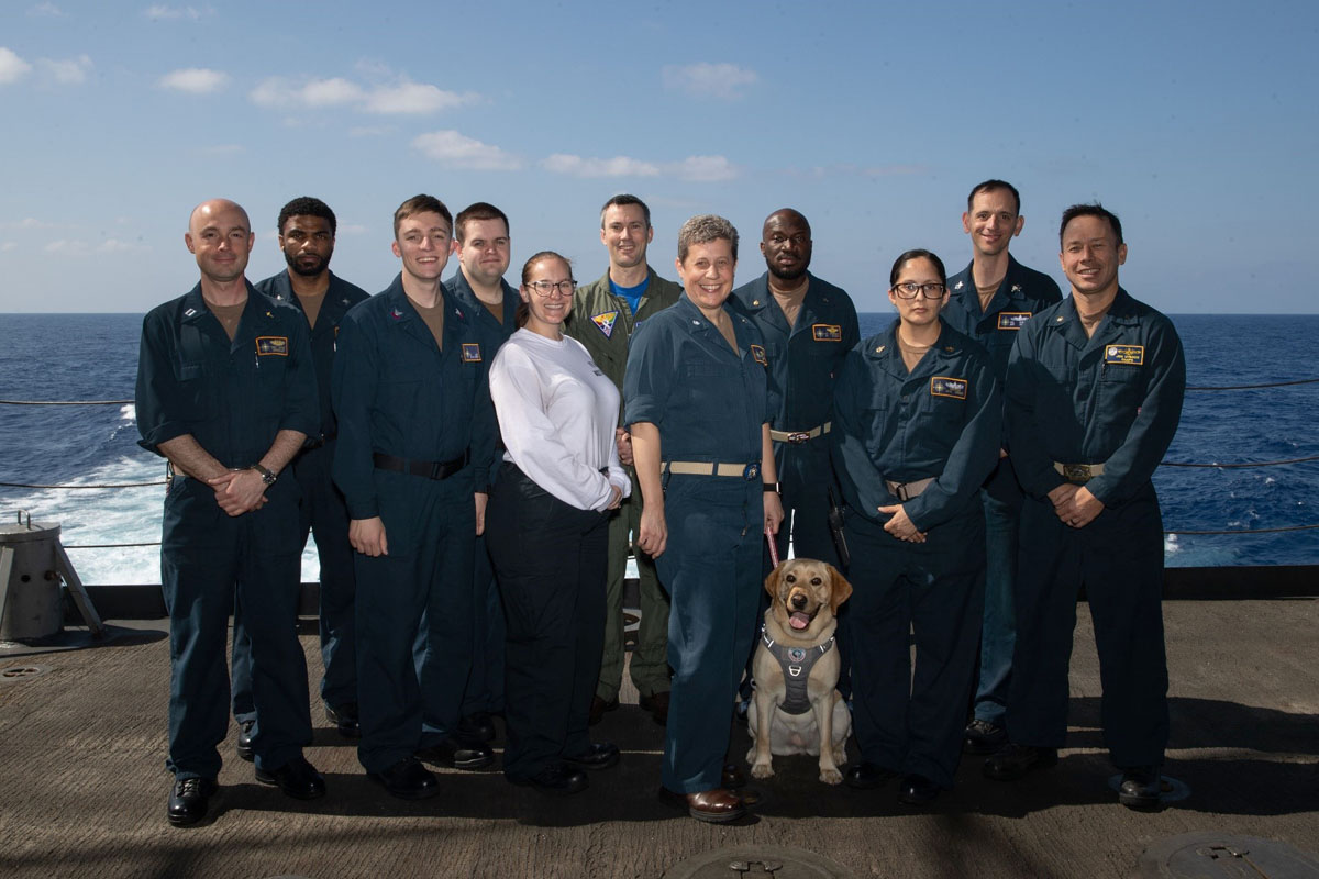U.S. Navy Commander Genevieve Clark (fifth from right) poses for a photo with the Command Religious Ministry Team aboard the USS Gerald R. Ford. Back row, left to right: Religious Program Specialist Arnold LaCour,  RPS Joseph Hunley, Lt. Commander Joshua Henderson (chaplain), Lt. Commander Mario Murphy (chaplain), unidentified chaplain. Front row, left to right: Lt. Commander Benjamin Pitre (chaplain), RPS Michael Day, Aviation Ordnanceman Abbie Chambers, Clark (chaplain), Sage (facility canine) and RPC Yanet Chavez Espinoza. Photo courtesy of Genevieve Clark.