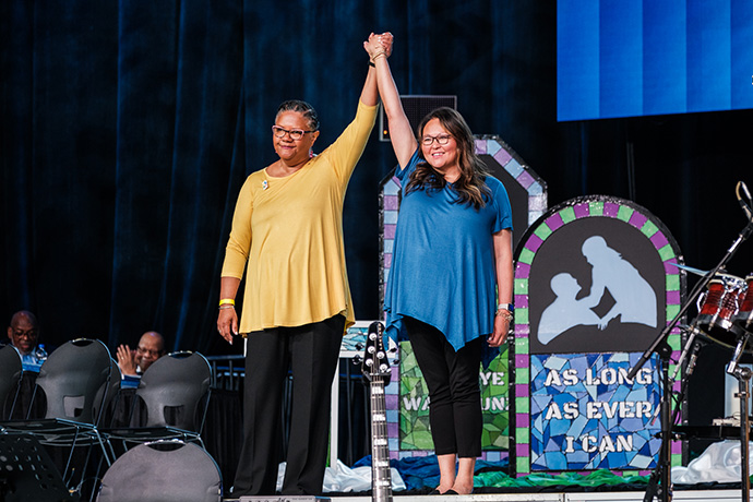 Sally Vonner, United Women in Faith top executive, and Bethany Amey Sutton, secretary of the group’s Executive Board, join hands on stage at the United Methodist General Conference in Charlotte, N.C., on April 29. Photo by Corbin Payne.