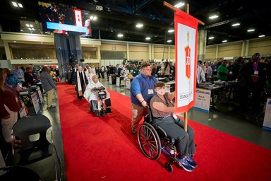 Sara Martin, her wheelchair pushed by Dan Levine, carries the banner of the Council of Bishops into the opening worship service of the United Methodist General Conference in Charlotte, N.C., on April 23. Behind them comes a procession of United Methodist bishops, headed by Bishop Forrest Stith on a motorized wheelchair. Photo by Paul Jeffrey, UM News.
