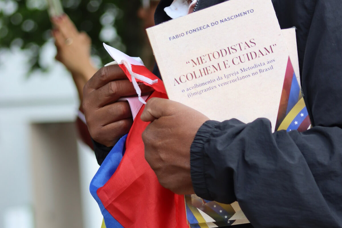 Durante conversa e caminhada com o autor, o Pastor Edson Cesar da Silva carrega o livro “Metodistas Acolhem e Cuidam” e a bandeira da Venezuela, país de onde o projeto atende a maior parte das pessoas acolhidas. Foto de Sara de Paula, para Notícias MU. 