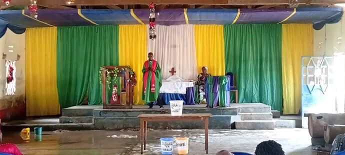 Le pasteur Kamukolobondo Kachoma Amisi Salomon (au centre) se tient sur la plate-forme de l'Église Méthodiste Unie Sayuni, décorée de rideaux colorés. Bien que l'église ait été lourdement endommagée par les inondations qui ont submergé l'intérieur, le pasteur et les fidèles se mobilisent ensemble pour reconstruire et soutenir les familles touchées. Photo avec l’aimable autorisation du Bureau de Gestion des Catastrophes de la Région Épiscopale du Tanganyika.