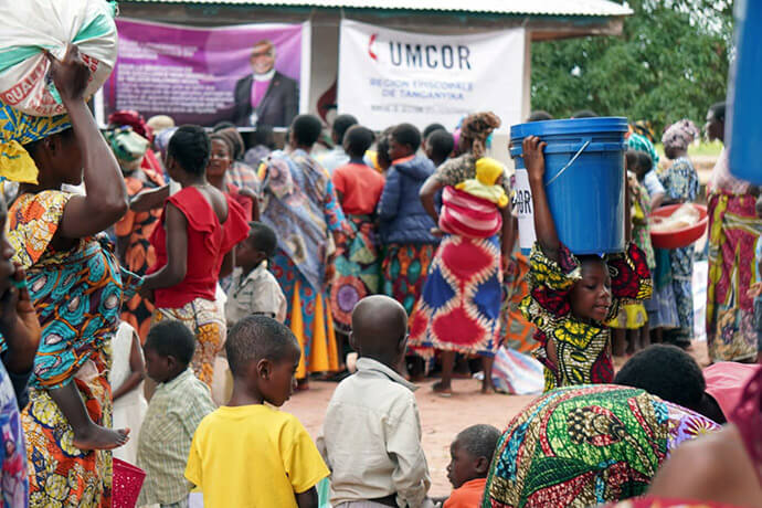 Une jeune fille porte un seau de provisions sur la tête au milieu d’une foule de bénéficiaires rassemblés devant des banderoles de l’UMCOR et de la région épiscopale de Tanganyika. La distribution de kits de secours vise à redonner leur dignité aux familles de Nyunzu, qui luttent pour survivre face à l’insécurité alimentaire et aux maladies résultant des récentes inondations et des conflits dans l’est de la RDC. Photo gracieusement fournie par le Bureau de gestion des catastrophes de la région épiscopale de Tanganyika.