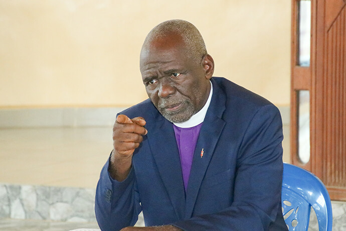 L'évêque Antoine Kalema Tambwe, de la Région Épiscopale du Congo Est, vêtu d'une aube blanche et d'une étole rouge, lève la main en signe de bénédiction tandis qu'il prie pour les épouses de pasteurs qui tiennent fièrement leurs certificats de formation, certaines portant des bébés. Cette prière de consécration a suivi une cérémonie de remise des certificats qui s'est tenue à Kindu, marquant l'engagement de ces femmes à devenir des leaders et des actrices clés du développement communautaire au sein de leurs paroisses rurales. Photo de Chadrack Tambwe Londe, UM News.