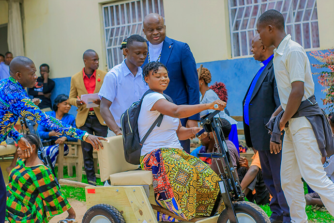Une jeune fille à mobilité réduite sourit alors qu’elle manœuvre pour la première fois son tricycle équipé de manivelles, sous le regard de l’Evêque Mande Muyombo (debout au centre) et d’autres responsables de l’Église. Ce projet vise à favoriser l’inclusion sociale et éducative en permettant aux jeunes et aux adultes de se rendre dignement sur leur lieu de travail ou d’études. Photo de Yamba Kisonga Barthélémy, UM News.