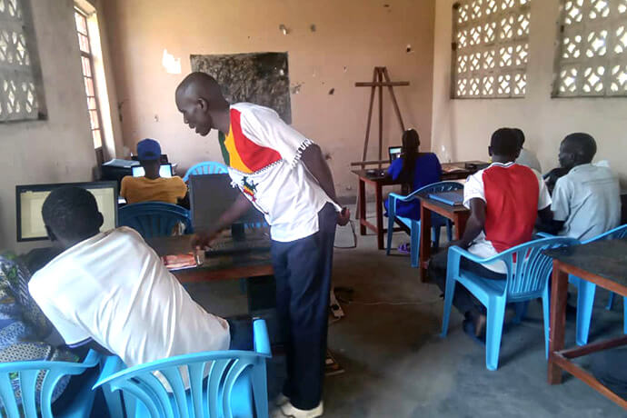Hakim Erezenious Kwenyo (standing) helps a student during a session of the Youth Digital Literacy Project at Ariaze United Methodist Church’s women’s training center in the Rhino Camp Settlement of Uganda’s Siripi Zone. The project is supported by The United Methodist Church’s South Sudan Children Ministry and the Holston Foundation. Photo by Mudomo Joseph, UM News. 